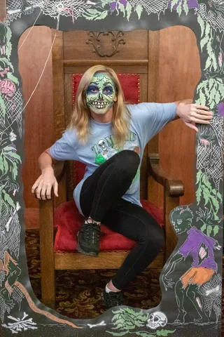A woman in green skull makeup and a Zombie Bike Parade t-shirt perches sideways in the carved lodge chair inside the painted zombie frame.