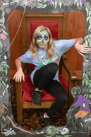 A woman in green skull face paint sits cross-legged in the carved lodge chair, one arm stretched along the painted frame beside her.