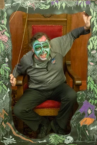 A man in green zombie makeup with a painted exposed brain throws one arm in the air from the carved lodge chair inside the painted frame.