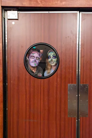 Two zombies in purple and green face paint peer through the round porthole window of a wooden kitchen door.