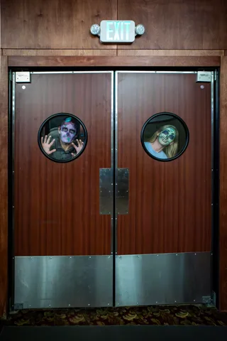 Two sets of swing doors below an EXIT sign, with a zombie pressing his hands to the glass in one porthole and a second zombie staring out of the other.