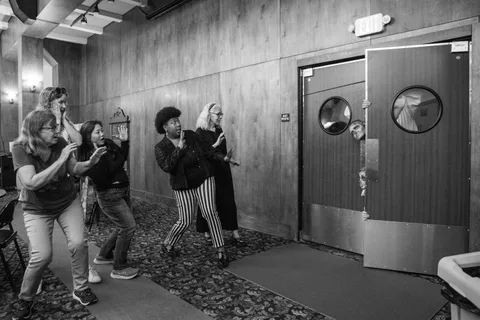 Black and white: five people in a panelled hallway recoil with their hands up as two zombies lean out from behind a swing door.