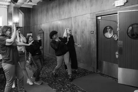 Black and white: a group backs away down the hallway while three zombies crowd the doorway, one framed in the porthole window.