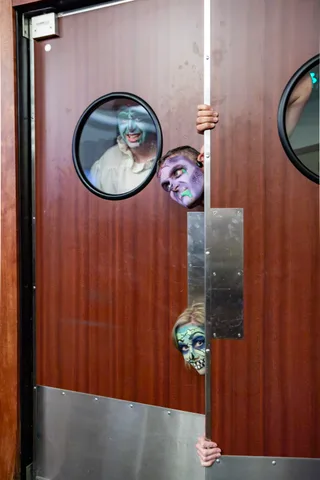 Three zombies squeeze around the edge of a swing door, one looking out through the porthole, one over the top and one peering around at waist height.