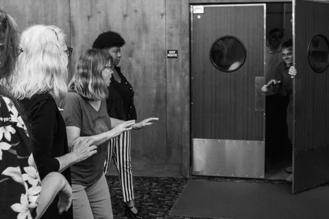 Black and white: four people stand back with their hands raised as a zombie pushes the swing door open from the other side.