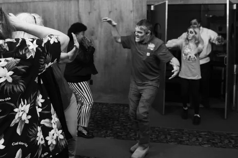 Black and white: zombies stride out through the open doors with their arms raised while the group in the hallway scatters.