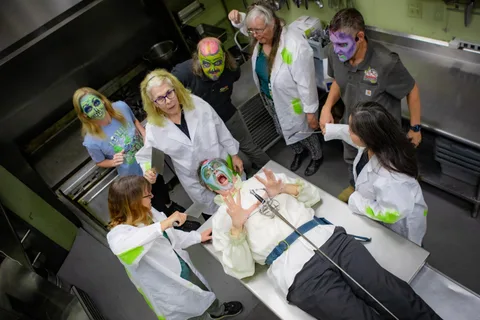 Looking down into the lodge kitchen: five people in white lab coats and zombie face paint crowd around a stainless steel counter where a zombie in a white shirt lies pretending to struggle.