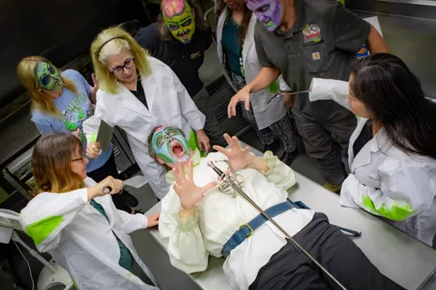 The mad scientists in lab coats lean over a zombie laid out on the stainless counter, one holding a cleaver and another a whisk, while he throws his hands up and yells.
