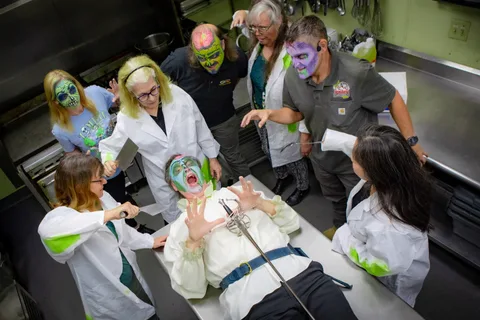 A wide view of the kitchen tableau: seven people in zombie makeup, most in white lab coats, surround a zombie lying on the counter with his hands raised.