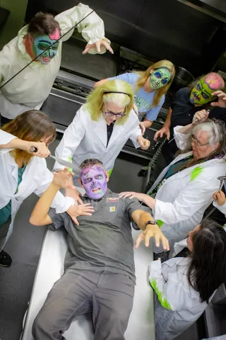 Looking down the length of the kitchen counter at a zombie lying flat with arms outstretched, ringed by lab-coated figures holding a knife and kitchen tongs.