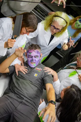 Close in on a zombie in purple face paint lying on the kitchen counter as two people in lab coats lean over him, one raising a cleaver.