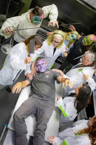 The full kitchen tableau from above: a zombie lies on the counter while eight others in lab coats and face paint reach in with knives, tongs and a skewer.