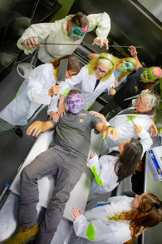 A vertical view of the kitchen scene, the zombie flat on the stainless counter and the lab-coated crowd closing in from every side.