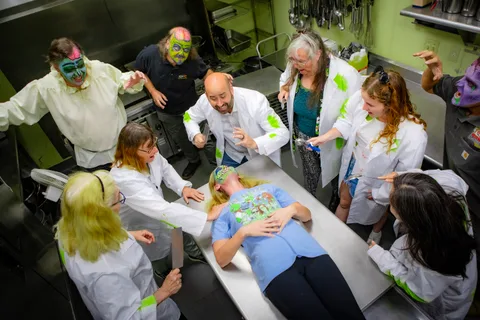 A second kitchen tableau: a zombie in a Zombie Bike Parade t-shirt lies on the stainless counter while nine people in lab coats and face paint lean in around her.