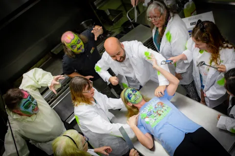 Close on the kitchen counter as a zombie in green face paint lies back, a cleaver held across her and the scientists leaning in overhead.