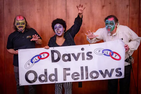 Three people in zombie makeup hold up a white Davis Odd Fellows banner against a wood-panelled wall, hands raised in a claw pose.