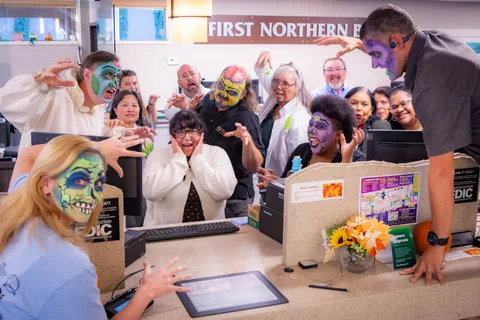 A wider frame of the same bank scene, a teller screaming with her hands to her cheeks while zombies in face paint reach over the counter around her.