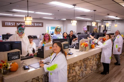 The full teller line at First Northern Bank, staff and zombies in face paint turning to smile at the camera along a stone-faced counter with pumpkins and autumn flowers.