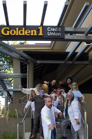 A group in zombie makeup, several in white lab coats, shambles down the entrance steps under the Golden 1 Credit Union sign.