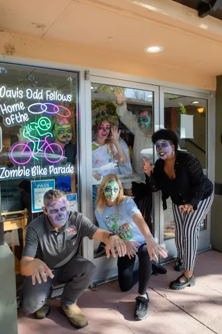 Four zombies crouch and lean outside the Odd Fellows Hall doors beside the neon sign reading Davis Odd Fellows, Home of the Zombie Bike Parade.