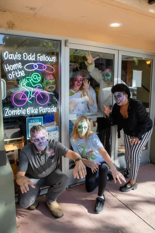 The same doorway from a step back: zombies posing outside the glass doors under the Zombie Bike Parade neon sign, with two more waving from inside.