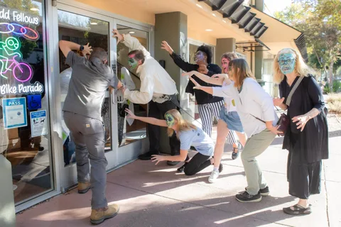 Zombies converge on the Odd Fellows Hall doors from the sidewalk while one man pulls the door open, the neon Zombie Bike Parade sign in the window beside them.