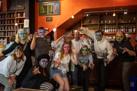 A dozen zombies pose with their hands up inside Mishka's Cafe, the espresso menu board and shelves of syrup bottles behind them.