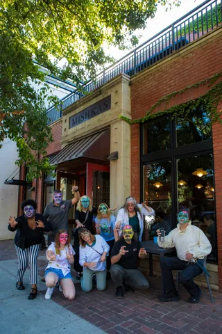 The zombie group poses on the brick sidewalk outside Mishka's Cafe, some kneeling, under the MISHKAS sign.