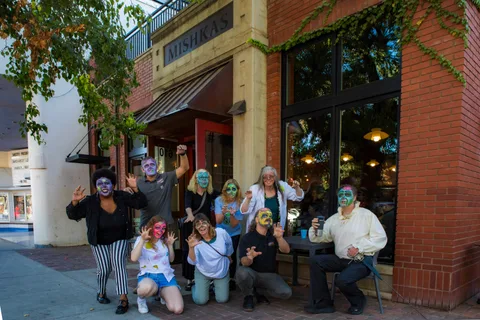 A wider frame outside Mishka's Cafe, the group in face paint waving and clawing at the camera along the storefront.
