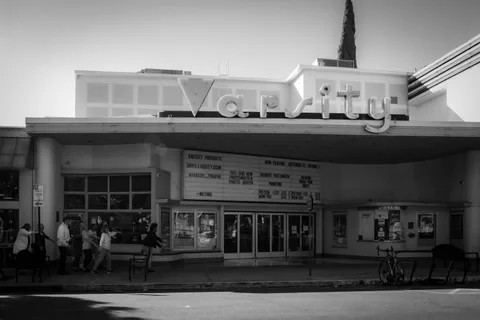 Black and white: the Varsity Theatre on a bright afternoon, its curved neon sign and marquee above the entrance, with a few people on the sidewalk.