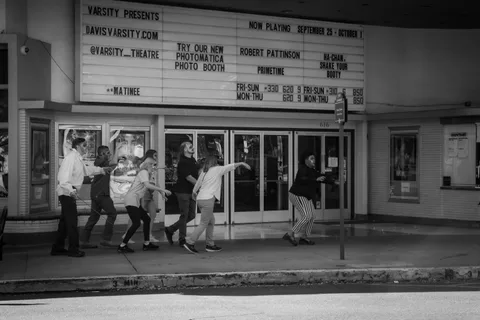 Black and white: a line of zombies shambles along the sidewalk past the Varsity Theatre doors under the marquee.