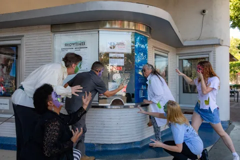 Zombies converge on the round Varsity Theatre ticket booth, one pretending to buy a ticket while the rest close in from both sides.