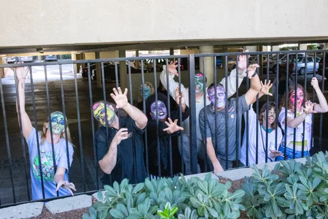 A dozen zombies crowd behind the parking garage railings with their arms thrust up through the bars, shrubs in the foreground.