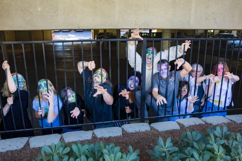 Twelve zombies line the parking garage railing, hands gripping the bars and reaching towards the camera, one shouting at the back.