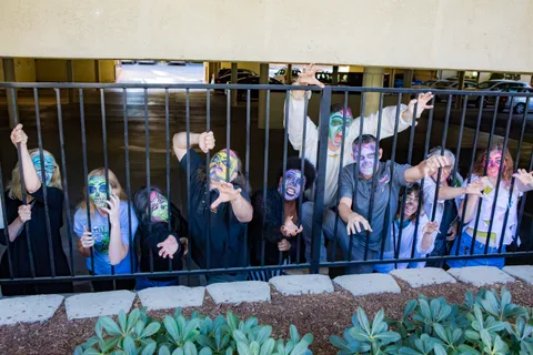 A wider frame of the railing line, zombies pressed against the bars with a concrete planter and shrubs in front.