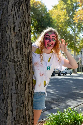 A zombie in a torn white t-shirt leans out from behind a tree trunk on a sunny street, waving and grinning at the camera.