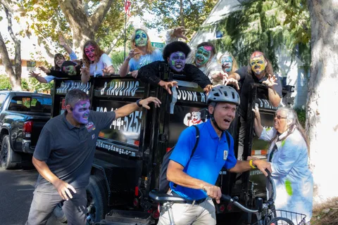 Close on the hauling trailer with zombies hanging over its rails, claws out, as a cyclist in a blue polo shirt rides past in front of them.