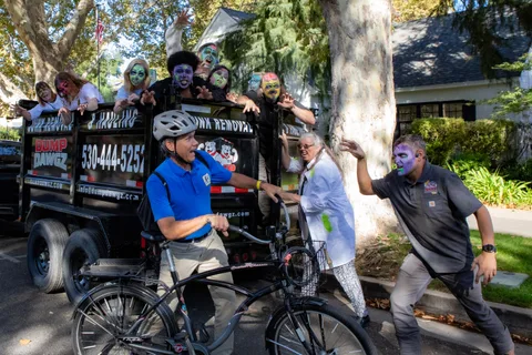 A cyclist in a helmet looks back over his shoulder as zombies lean out of the Dump Dawgz hauling trailer and two more close in from the kerb.