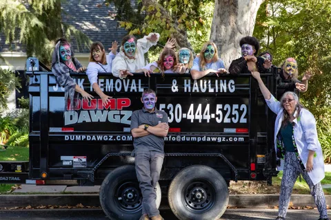Ten zombies pose along the top rail of the black Dump Dawgz junk removal trailer, one standing in front with his arms folded.