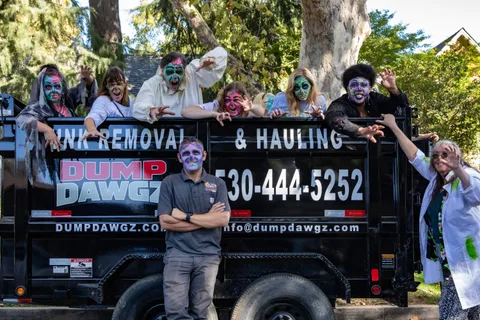 A group in zombie face paint lean out of a black Dump Dawgz dump trailer with clawed hands while a man in a work polo stands calmly in front, arms folded.