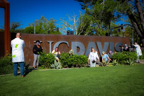 A man in a green-splattered lab coat photographs the zombie group as they pose along the UC DAVIS entry sign.