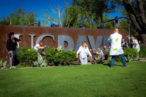 The zombie group shuffles toward a man in a splattered lab coat in front of the UC DAVIS sign, arms outstretched.