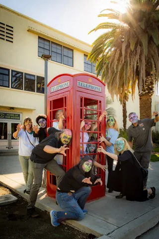 Zombies crowd around and inside a red British telephone box outside a campus building, hands raised, a palm tree and sun flare overhead.