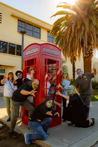 The group clusters around a red telephone box in the afternoon sun, several pressed against the glass inside and others clawing at the doors.
