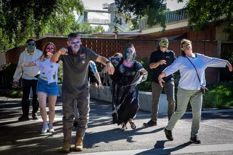 The group shuffles down a campus path in a loose line, arms hanging forward in a slow zombie walk.
