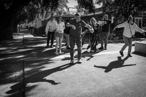 A black and white frame of the zombie walk along a tree-shaded campus path, long shadows stretching across the pavement.