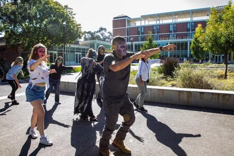 Zombies lurch across the plaza in front of the Memorial Union, a man in front leading with both arms thrown wide.