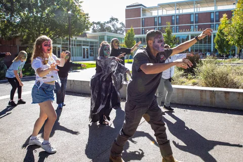 The group advances past the Memorial Union in bright sun, hands raised and faces painted green and purple.