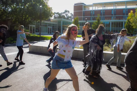 A woman in a torn and stained t-shirt roars at the camera mid-stride while the rest of the group shuffles behind her near the Memorial Union.