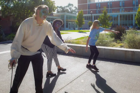 A man in a ruffled shirt carrying a thin sword leads two others in a slow zombie walk across the sunlit plaza.